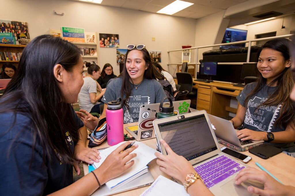 Smiling students sit at a table and work on their laptops.